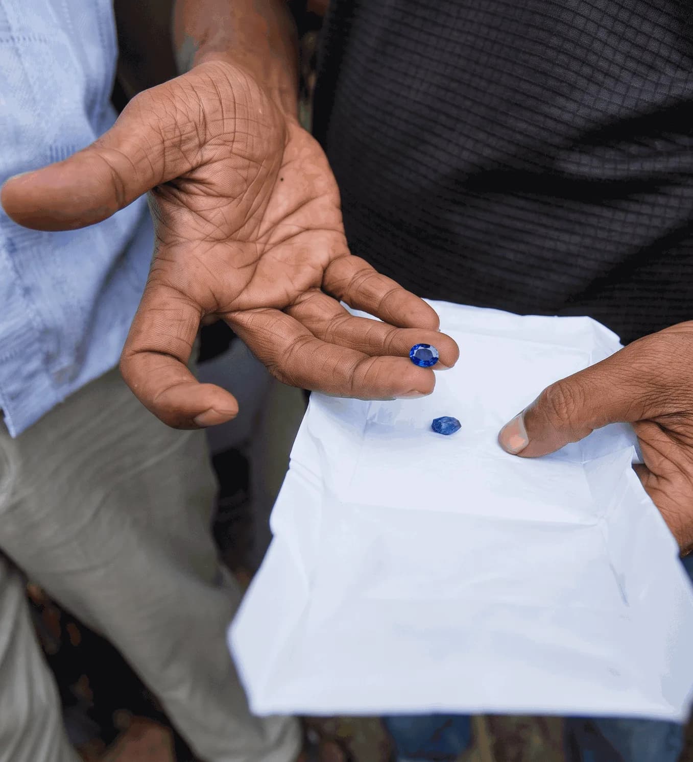 A cut blue Ceylon sapphire held at the fingertips over a white gem paper, with a rough blue sapphire lying on the paper